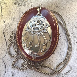 May include: Silver-toned oval locket pendant with intricate floral and scrollwork design, suspended from a silver chain. The locket rests on a red leather case with gold trim. A classic piece of jewelry.