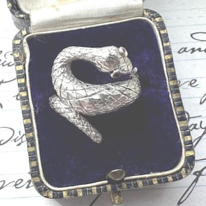 May include: A silver snake ring with red gemstone eyes, coiled inside a vintage blue velvet jewelry box. The ring is detailed with scales and is a unique piece of jewelry.