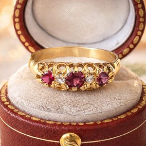 May include: A gold ring with five gemstones, set in a decorative band. The ring features three red gemstones and two smaller white gemstones. The ring is displayed inside a red jewelry box with a cream-colored interior.