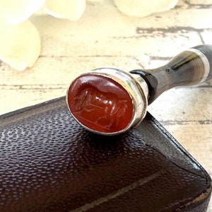 May include: A silver and agate handled wax seal with a red carnelian stone featuring a carved design. The seal is resting on a brown leather box.