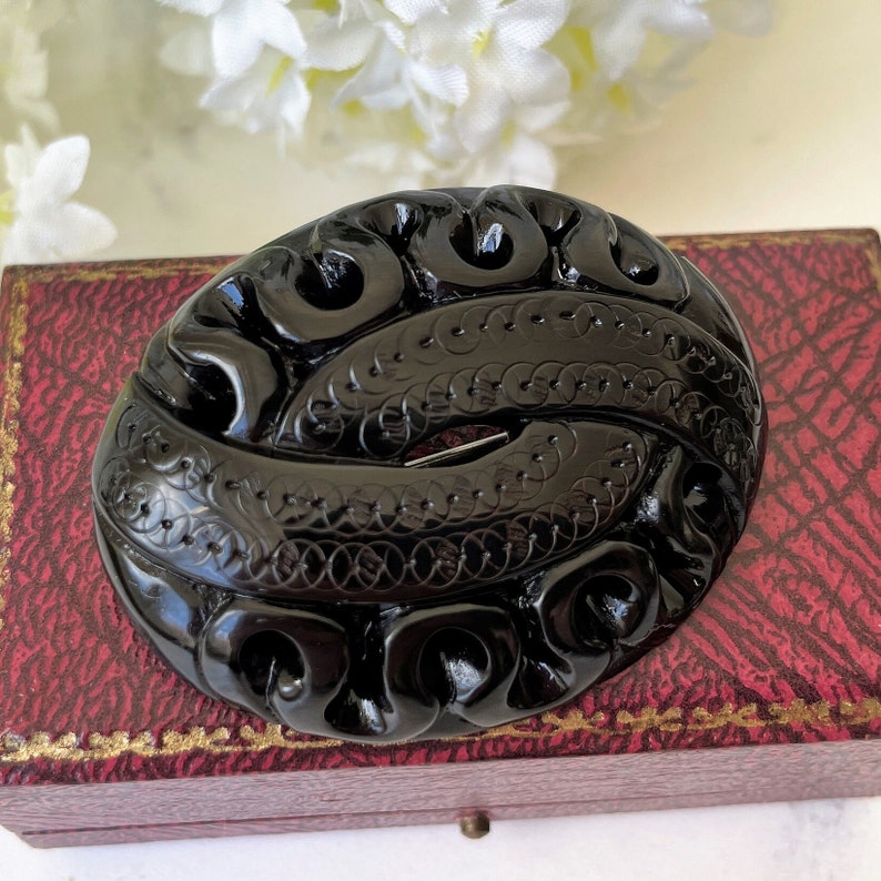 May include: A black carved jet brooch with an intricate design. The brooch is oval shaped and has a shiny finish.