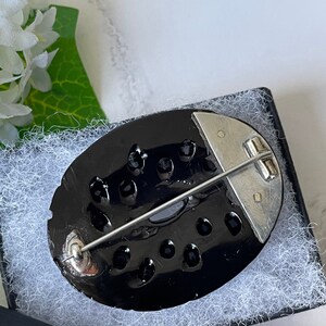 May include: A black oval brooch with a silver pin and clasp. The brooch is decorated with a pattern of small black circles.