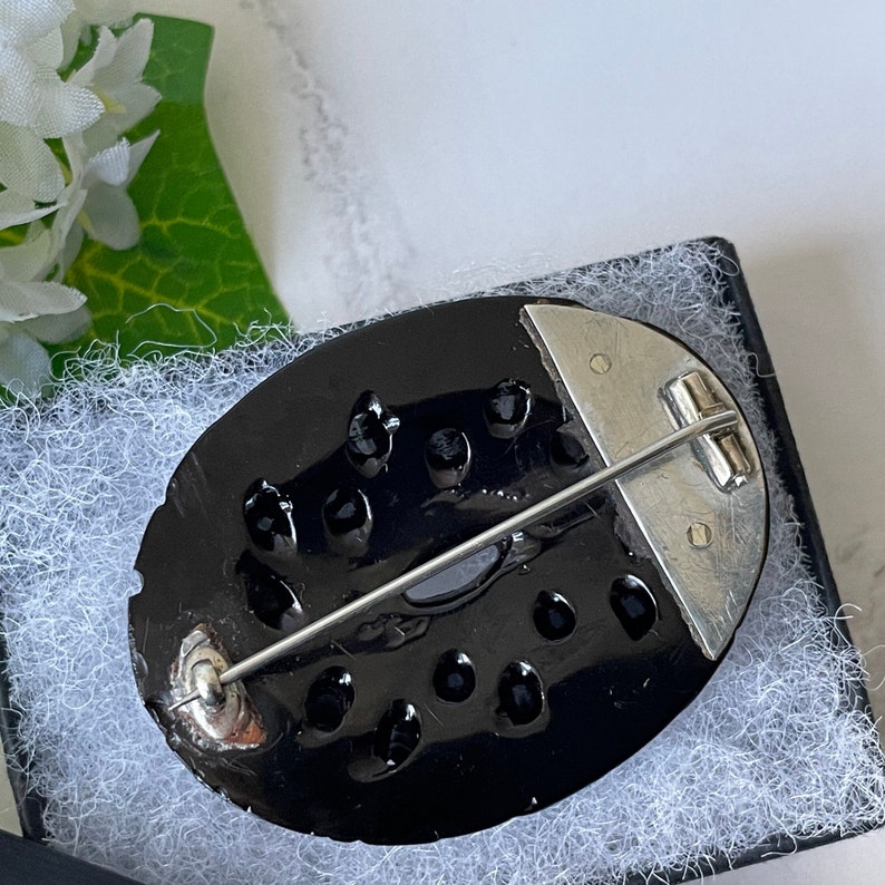 May include: A black oval brooch with a silver pin and clasp. The brooch is decorated with a pattern of small black circles.