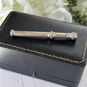 May include: A silver-toned cylindrical needle case featuring a fine cross-hatched texture along the body, accented by two decorative bands with intricate floral scrollwork engravings at the center and the ends of the elongated metal tube.