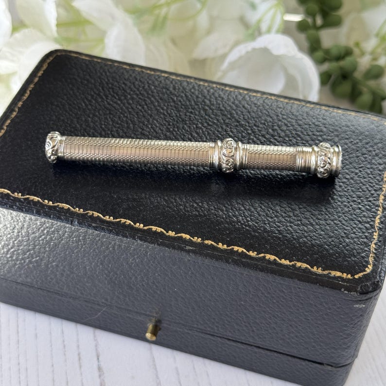 May include: A silver-toned cylindrical needle case featuring a fine cross-hatched texture along the body, accented by two decorative bands with intricate floral scrollwork engravings at the center and the ends of the elongated metal tube.