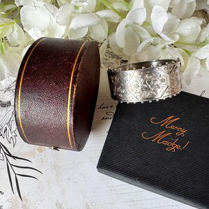 May include: A silver bangle bracelet with an intricate floral design. The bracelet is in a brown box with a gold trim and a black box with the words "Mercy Madge!" in gold lettering.