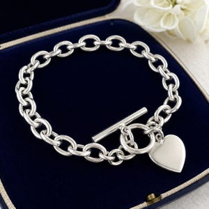 Könnte beinhalten: Ein silberfarbenes Armband mit Knebelverschluss und Herzanhänger. Das Armband besteht aus ovalen Kettengliedern. Der Herzanhänger ist glatt und poliert. Das Armband wird in einer blauen Samt-Schmuckschatulle präsentiert.