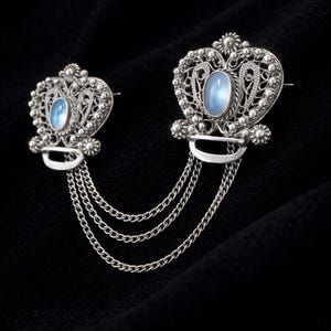 Puede incluir: Dos broches plateados unidos por cadenas. Cada broche presenta una piedra ovalada azul claro engastada en un diseño de filigrana ornamentado. Los broches tienen forma de corazón con elementos florales decorativos.