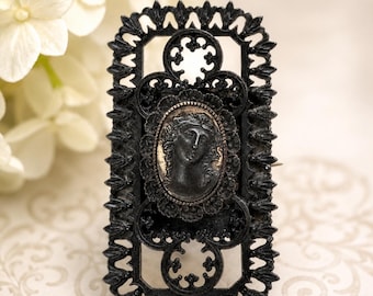 May include: A rectangular black brooch featuring an intricate openwork metal frame with a pointed edge design. At the center, an oval medallion displays a raised relief profile of a human face in a dark finish with subtle metallic highlights.