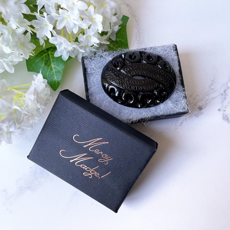 May include: A black oval brooch with a carved design, in a black box with the text "Mercy Madge!" in gold lettering.