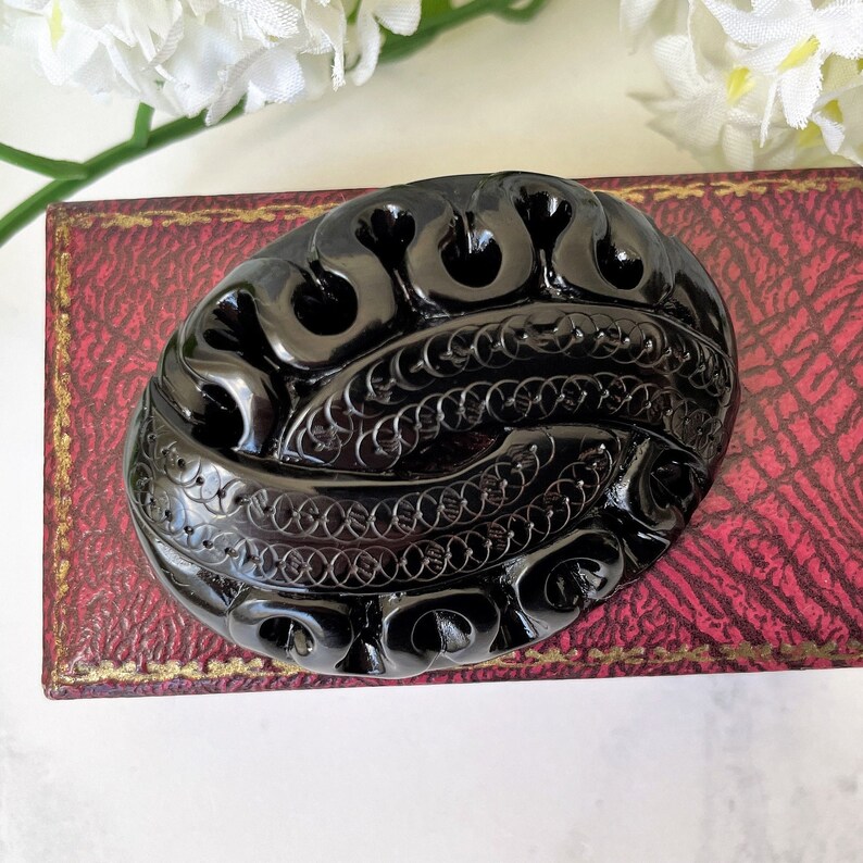 May include: A black carved oval brooch with intricate designs. The brooch is made of a shiny, smooth material and has a detailed pattern of swirls and circles.
