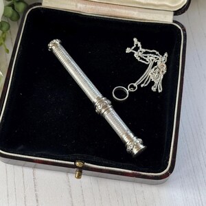 May include: A silver cylindrical needle case with intricate cross-hatched engraving and decorative filigree end caps, accompanied by a delicate silver chain featuring a small circular ring attachment, all presented inside a dark velvet-lined box.