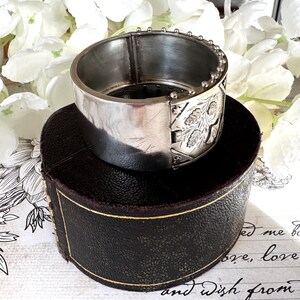 May include: A silver hinged bangle bracelet with an engraved floral design. The bracelet is sitting on a brown box with a gold trim.