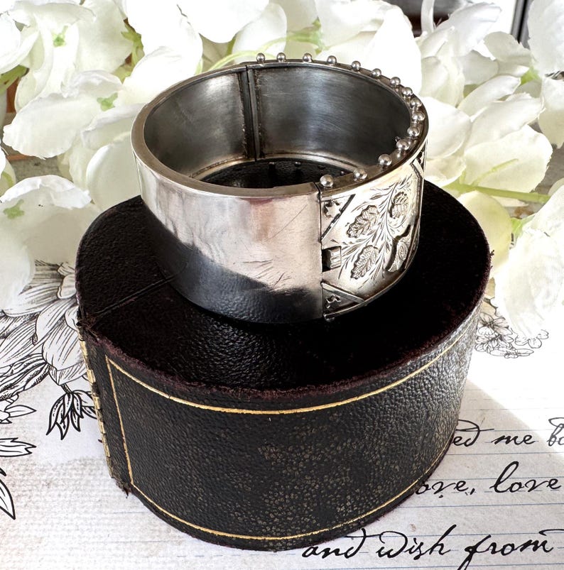 May include: A silver hinged bangle bracelet with an engraved floral design. The bracelet is sitting on a brown box with a gold trim.