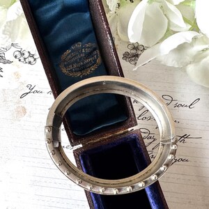 May include: A silver bangle bracelet with a hinged clasp and a row of small beads around the edge. The bracelet is in a blue velvet box with a label that reads "Bressey & Son Watchmakers 326 High Street Stratford E".