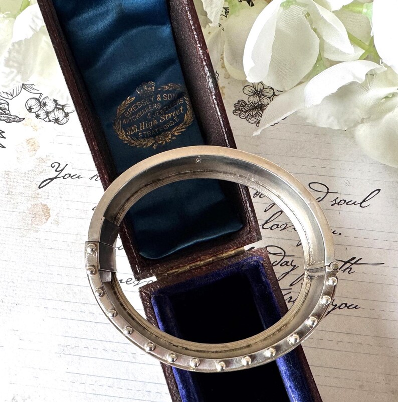 May include: A silver bangle bracelet with a hinged clasp and a row of small beads around the edge. The bracelet is in a blue velvet box with a label that reads "Bressey & Son Watchmakers 326 High Street Stratford E".