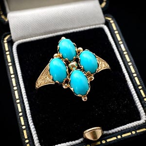 May include: A gold ring with four oval turquoise stones arranged in a cross shape. The ring has intricate floral detailing on the band and is set in a black jewelry box.