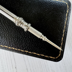 May include: A silver mechanical pencil featuring an intricate engine-turned crosshatch pattern on the barrel, accented by decorative embossed scrollwork bands near the center and a tapered tip, resting against a black leather case with gold stitching.