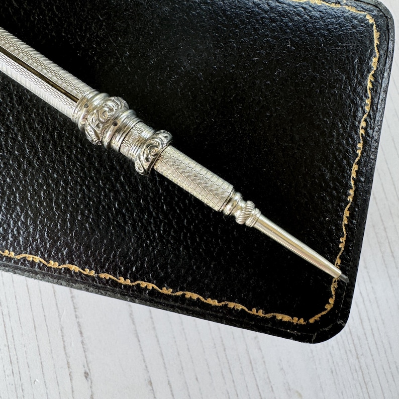 May include: A silver mechanical pencil featuring an intricate engine-turned crosshatch pattern on the barrel, accented by decorative embossed scrollwork bands near the center and a tapered tip, resting against a black leather case with gold stitching.