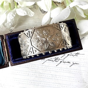 May include: A silver bracelet with an intricate floral design. The bracelet is engraved with a pattern of roses and leaves. The bracelet is in a small blue velvet box.