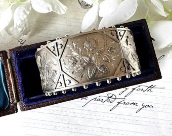 Puede incluir: Un brazalete de plata con un intrincado diseño floral. El brazalete está grabado con un patrón de rosas y hojas. El brazalete está en una pequeña caja de terciopelo azul.