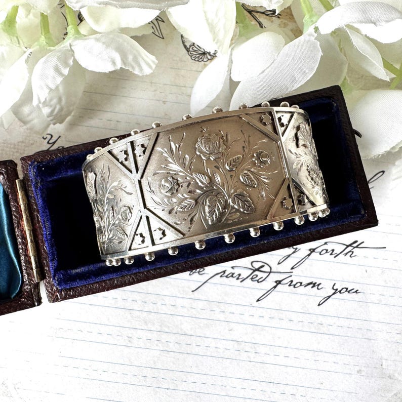 May include: A silver bracelet with an intricate floral design. The bracelet is engraved with a pattern of roses and leaves. The bracelet is in a small blue velvet box.