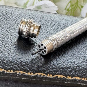 May include: A silver mechanical pencil with a textured, cross-hatched barrel and a removable cap featuring an ornate, embossed floral pattern. The pencil tip displays a circular arrangement of six small holes with a thin piece of graphite lead.