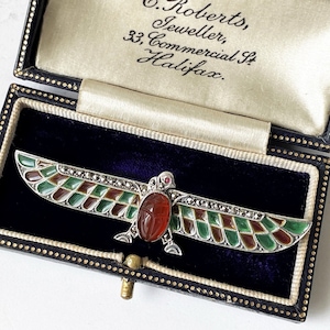 May include: A silver and enamel brooch in the shape of a winged scarab beetle. The wings are green, brown, and gold, and the scarab is red. The brooch is in a black velvet box with a white satin lining. The box has a label that reads "C. Roberts, Jeweller, 33, Commercial St, Halifax."