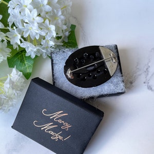 May include: A black oval brooch with a silver pin and a black and white design. The brooch is in a black box with the words "Mercy & Madge!" written in gold.