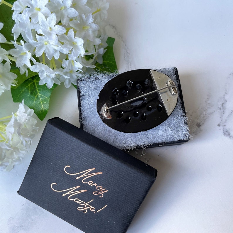 May include: A black oval brooch with a silver pin and a black and white design. The brooch is in a black box with the words "Mercy & Madge!" written in gold.