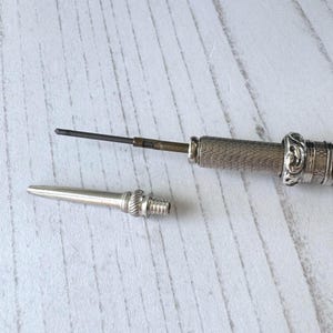 May include: A silver mechanical pencil with a textured, cross-hatched barrel and an ornate, embossed ring near the tip. The pointed metal cap is detached, revealing a thin, dark graphite lead extending from the internal brass mechanism.
