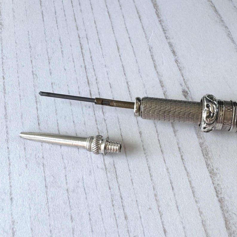 May include: A silver mechanical pencil with a textured, cross-hatched barrel and an ornate, embossed ring near the tip. The pointed metal cap is detached, revealing a thin, dark graphite lead extending from the internal brass mechanism.