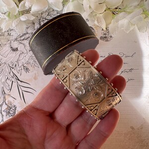 May include: A silver bangle bracelet with an intricate floral design. The bracelet is engraved with a repeating pattern of flowers and leaves. The bracelet is held in a hand.
