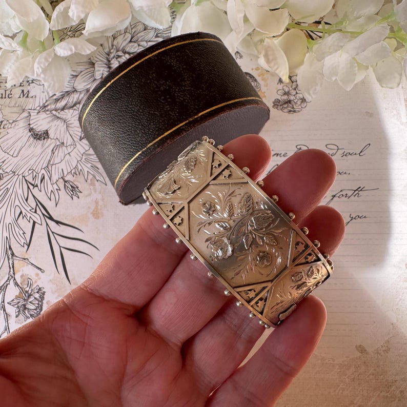 May include: A silver bangle bracelet with an intricate floral design. The bracelet is engraved with a repeating pattern of flowers and leaves. The bracelet is held in a hand.