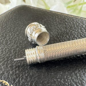 May include: A silver mechanical pencil with a fine wavy line texture on the barrel and a screw-off cap featuring a floral embossed pattern. The pencil tip is unscrewed, revealing a thin piece of dark graphite lead extending from the internal mechanism.