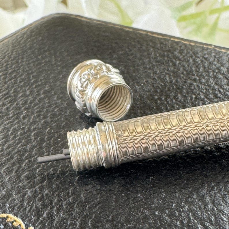 May include: A silver mechanical pencil with a fine wavy line texture on the barrel and a screw-off cap featuring a floral embossed pattern. The pencil tip is unscrewed, revealing a thin piece of dark graphite lead extending from the internal mechanism.