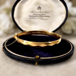 May include: A gold-toned hinged bangle bracelet with a faceted geometric surface, featuring a safety chain and a clasp. The bracelet rests inside an oval-shaped jewellery box with a dark blue velvet interior and a cream-coloured silk lid lining.