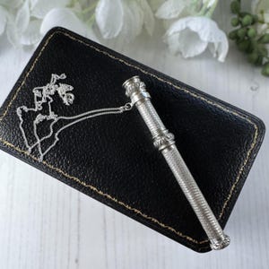 May include: A silver cylindrical needle case with a textured, cross-hatched pattern and decorative bands at the ends. A thin silver chain is attached to the top cap, extending across a black rectangular case with visible gold-colored stitching.