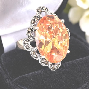 Puede incluir: Anillo de plata con una gran gema naranja de talla ovalada. La gema está engastada en un engaste de plata decorativo con piedras más pequeñas y oscuras. El anillo se encuentra sobre un fondo de terciopelo negro.