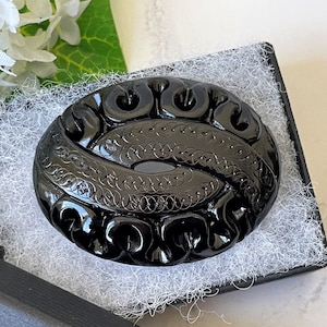 May include: A black carved glass brooch with an intricate design of swirls and circles. The brooch is oval shaped and has a high gloss finish.