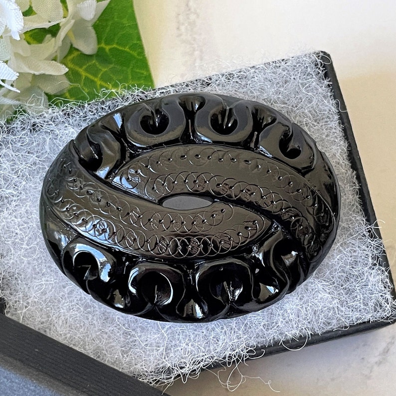 May include: A black carved glass brooch with an intricate design of swirls and circles. The brooch is oval shaped and has a high gloss finish.