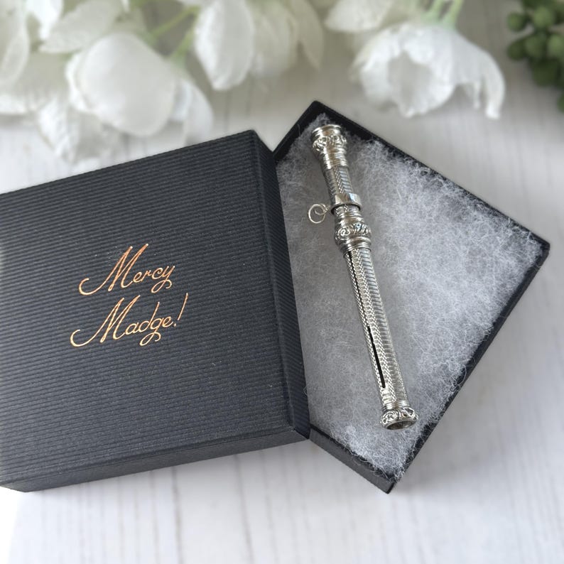 May include: A silver-toned cylindrical metal case with intricate engraved detailing and a small attached ring, resting inside a black square gift box. The lid of the box features the gold-colored text Mercy Madge! printed in a cursive script.
