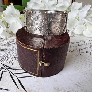 May include: A silver bangle bracelet with an intricate floral design and a beaded edge. The bracelet is in a brown leather box.