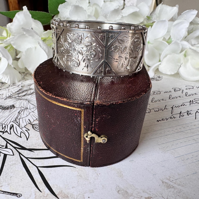 May include: A silver bangle bracelet with an intricate floral design and a beaded edge. The bracelet is in a brown leather box.