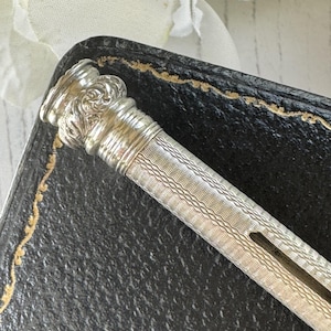 May include: A silver mechanical pencil featuring a cylindrical body with a fine, wavy guilloche pattern and a decorative, embossed floral motif at the top cap. The writing instrument has a slim, elongated shape with a visible side slot for the lead.