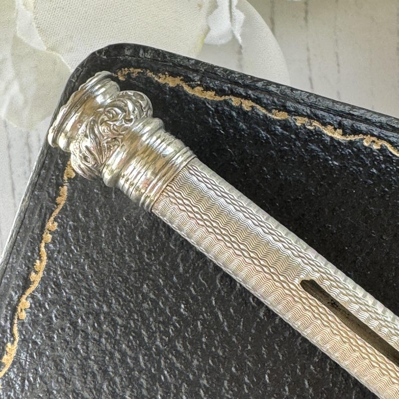May include: A silver mechanical pencil featuring a cylindrical body with a fine, wavy guilloche pattern and a decorative, embossed floral motif at the top cap. The writing instrument has a slim, elongated shape with a visible side slot for the lead.
