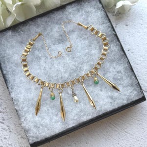 May include: A gold-toned necklace with a chain-link design. The necklace features several dangling pendants, including elongated gold shapes, small green beads, and a pearl-like charm. The necklace is displayed in a black box with a gray interior.