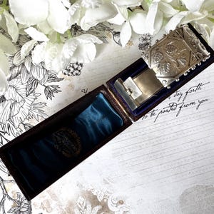 May include: A silver locket with an ornate floral design, in a dark brown velvet lined box. The box is open and the locket is resting on a blue velvet cushion. The text "to be parted from you" is written on a piece of paper in the background.