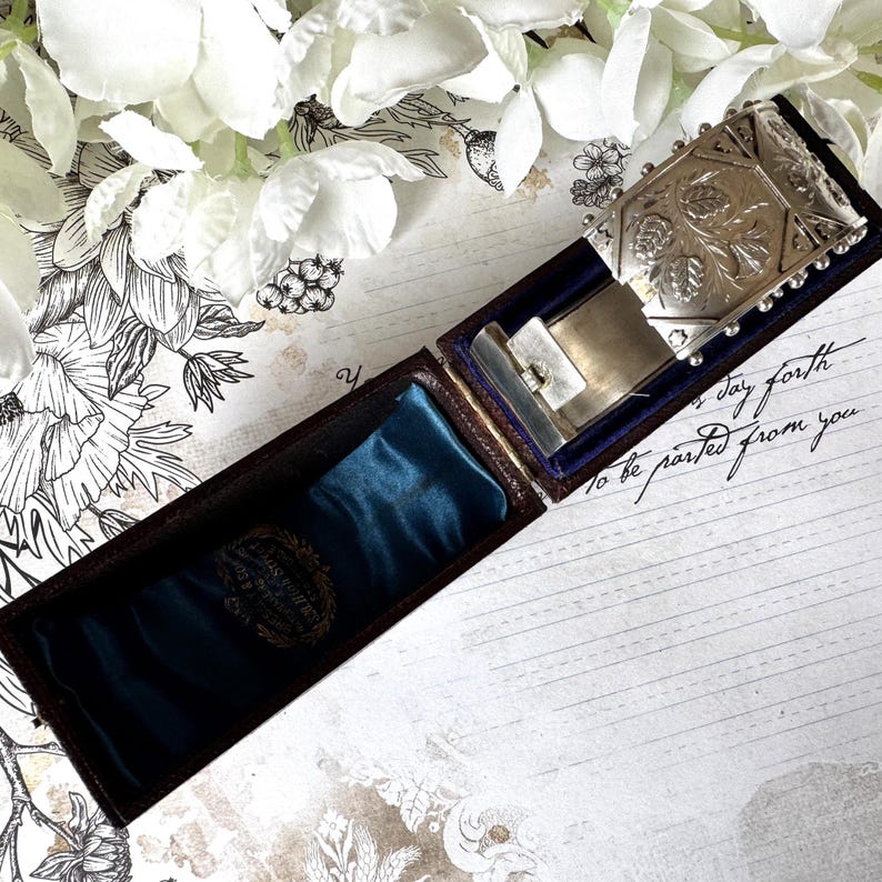 May include: A silver locket with an ornate floral design, in a dark brown velvet lined box. The box is open and the locket is resting on a blue velvet cushion. The text "to be parted from you" is written on a piece of paper in the background.