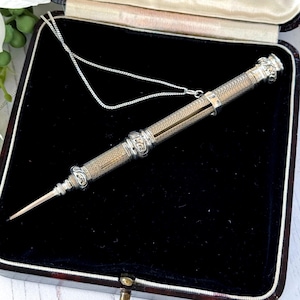 May include: A silver mechanical pencil with a textured, cross-hatched barrel and ornate, engraved metal bands. The pencil features a slender, pointed tip and is attached to a delicate, thin silver chain, presented inside a dark-lined case.